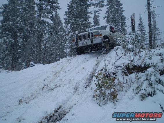 White 1992 Ford Bronco driving up a snowy hill with trees in the background
