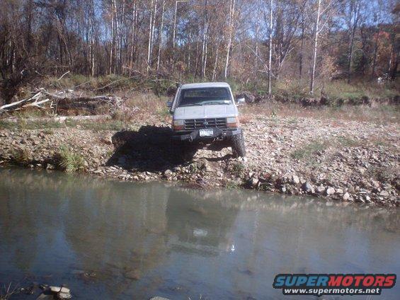 Gray Ford Ranger parked on rocky shore next to water, front view