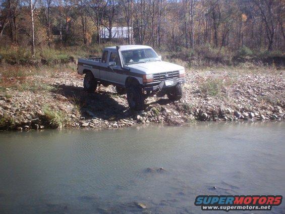 White 1990 Ford Ranger pickup truck with lift kit parked on a rocky river bank
