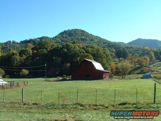 Rural landscape with red barn, green fields, trees, and mountains under a clear blue sky