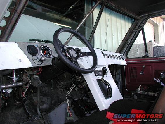 Customized interior of a vehicle, showing a racing steering wheel, aftermarket gauges, and a floor shifter. Roll cage visible