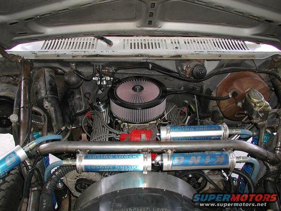 Engine bay with custom racing shocks, large air filter, and V8 engine components. Blue and silver shocks are visible