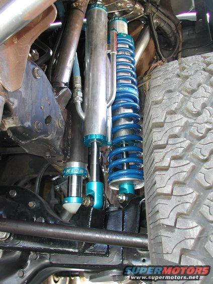Close-up of blue coilover shocks and silver reservoir shocks on a vehicle's suspension, with a tire visible