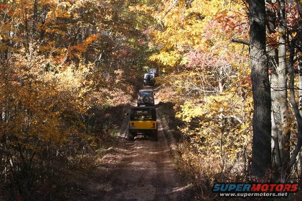 Yellow Jeep Cherokee leads a line of off-road vehicles on a dirt path through autumn woods. 1992 Jeep Cherokee