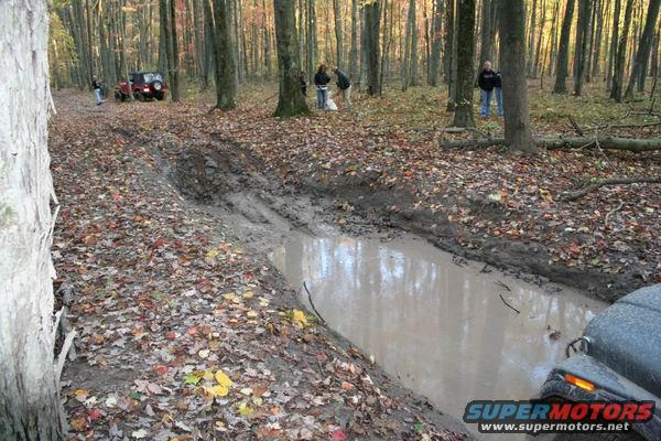 Red Jeep Wrangler on a muddy forest trail with people nearby