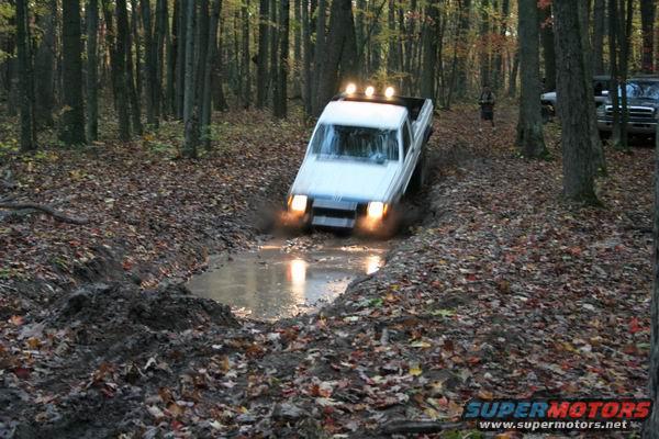 White pickup truck driving through mud, front view. Headlights on, splashing water