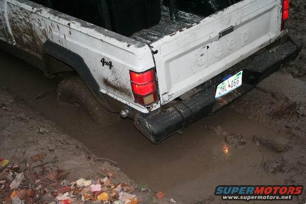 White Jeep Comanche pickup truck rear end stuck deep in mud, one tire submerged