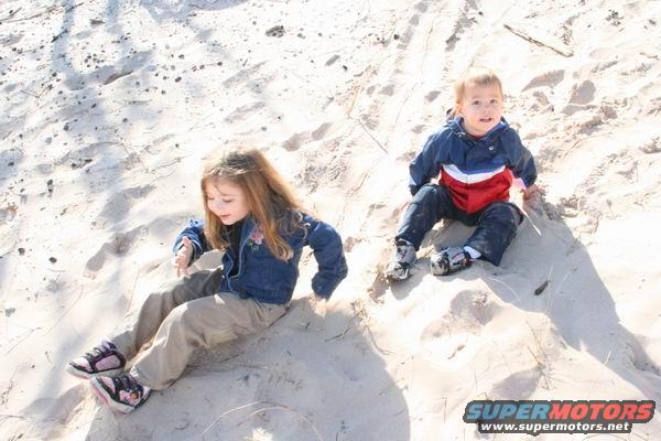 Children sitting on a sand dune, one girl in a denim jacket, one boy in a red, white, and blue jacket. A website URL is visible at the bottom right