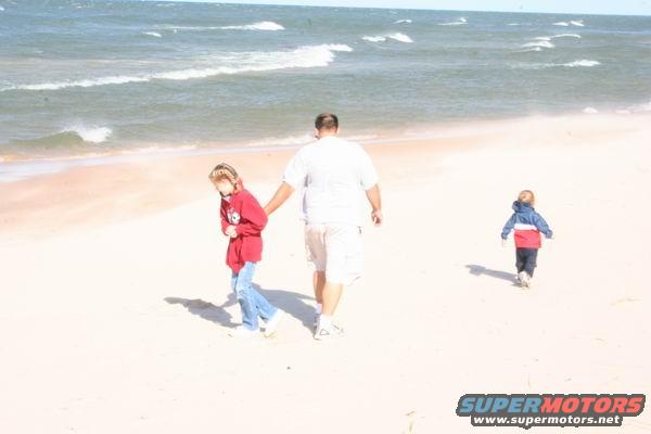 Man holding a child's hand on a sandy beach with the ocean in the background. Another child walks away from them towards the water