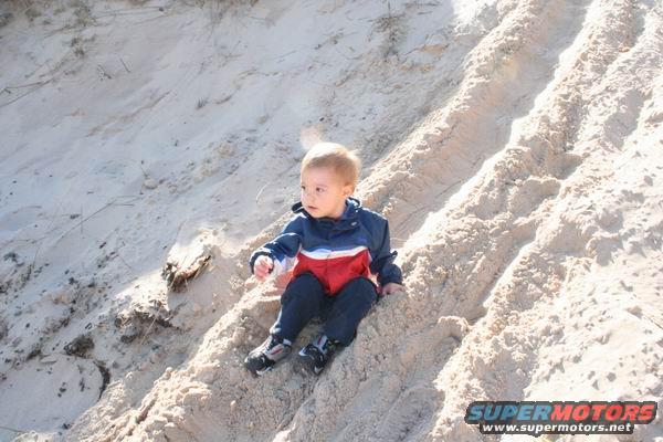 Child in blue, red, and white jacket sits on a sandy slope with tire tracks