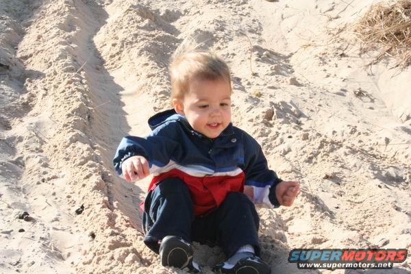 Toddler in blue, red, and white jacket sits in sand with tire tracks behind