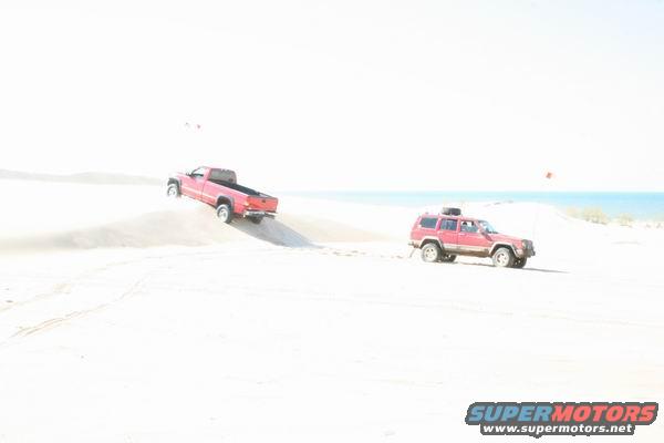 Red pickup truck driving up a sand dune, red SUV parked on sand nearby. Ocean in background