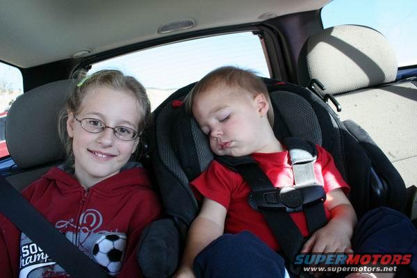 Girl with glasses smiles next to a sleeping toddler in car seats