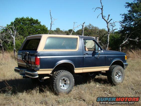 Blue and tan 1989 Ford Bronco with lifted suspension parked in a grassy, wooded area