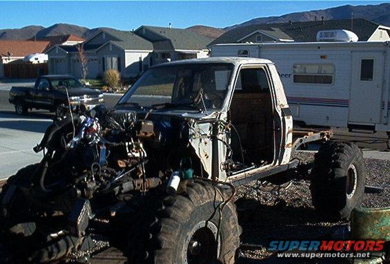 Off-road truck chassis with engine exposed, large tires, and a trailer in the background. Another truck is parked nearby