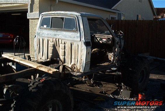 Pickup truck frame with cab, missing body panels and engine. Big tires on the chassis