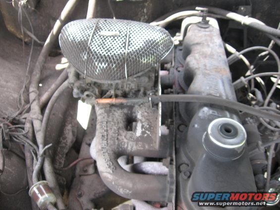 Close-up of a vehicle engine bay showing a carburetor, air cleaner, and exhaust manifold. Some wiring and hoses are visible