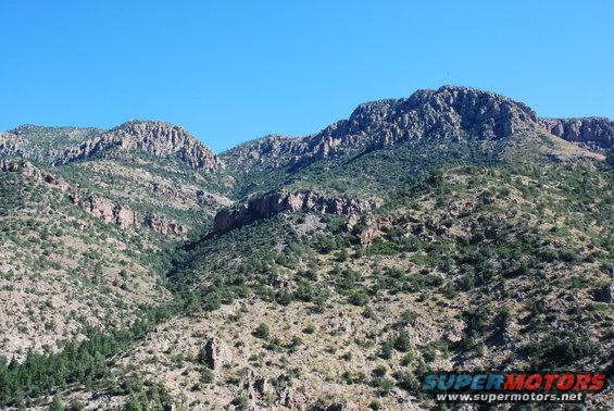 Rocky mountains with scattered trees under a clear blue sky. Watermark reads SUPER MOTORS