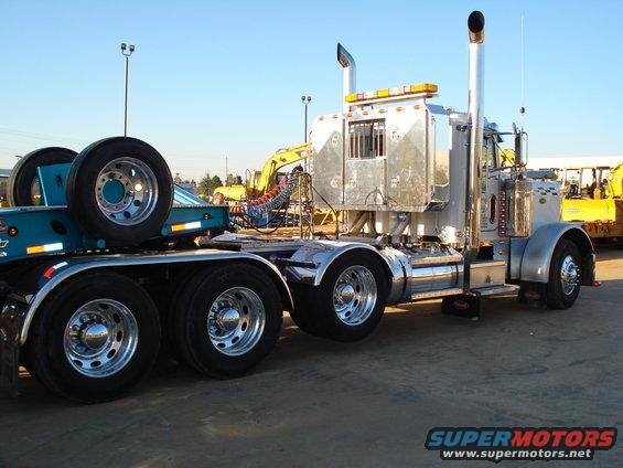 Silver semi-truck with dual rear wheels and a trailer carrying a spare tire, with a yellow excavator in the background