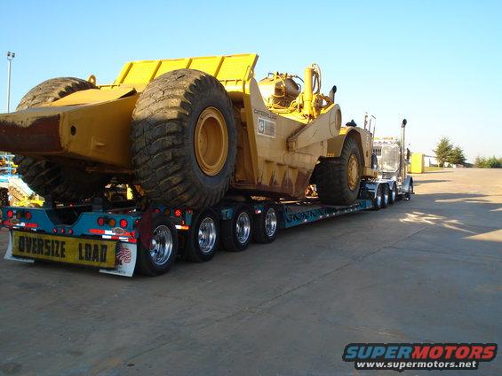 Yellow Caterpillar wheel tractor-scraper loaded on a multi-axle flatbed trailer with 'OVERSIZE LOAD' sign