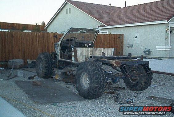 Custom off-road vehicle chassis with large tires, stripped body, and exposed interior, viewed from the front-right