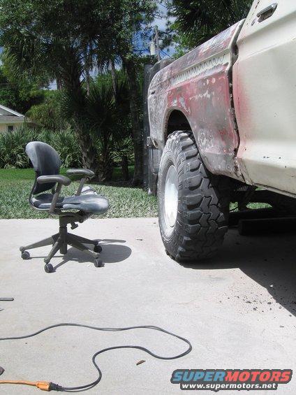1979 Ford F-150 side view showing rear fender and large off-road tire. Body is in primer with some red showing through. Office chair sits nearby
