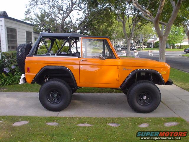 Orange 1977 Ford Bronco with black soft top and roll cage, parked on a sidewalk