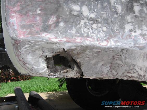 Vehicle body panel with extensive rust damage and body filler, showing a hole in the lower section. A tire is visible