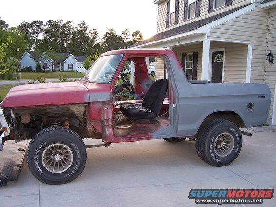 Side view of a red and gray Ford Bronco undergoing restoration, missing its front fenders and hood