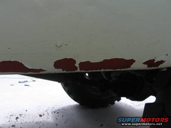 White truck lower body panel with chipped red paint, showing tire and suspension components