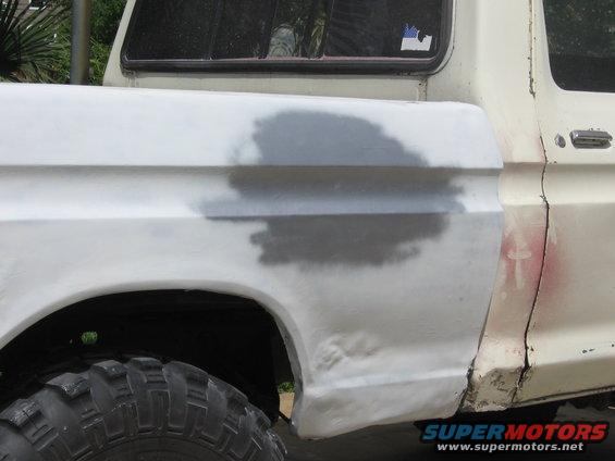 Side view of a white truck's rear fender and bed, showing body filler and primer. Tire visible