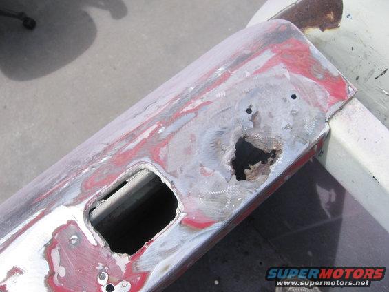 Close-up of a damaged and rusted vehicle panel, showing areas of red primer and exposed metal. There's a large hole and a smaller rectangular cutout