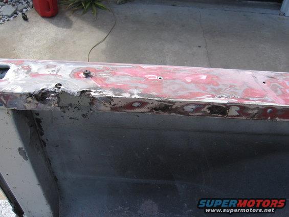 1979 Ford F-150 truck bed rail with rust and body filler, showing red paint underneath. A screw is visible