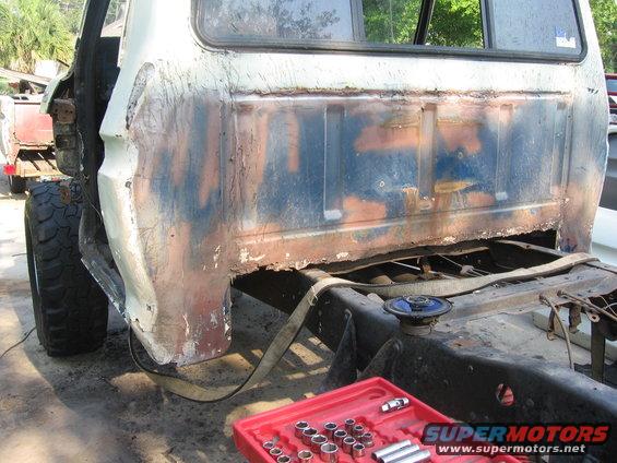 Truck bed with significant rust and damage, showing exposed frame and a socket set. Visible tire and part of a trailer