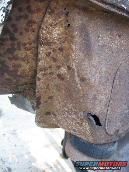 Heavily rusted metal panel with holes, showing suspension components below