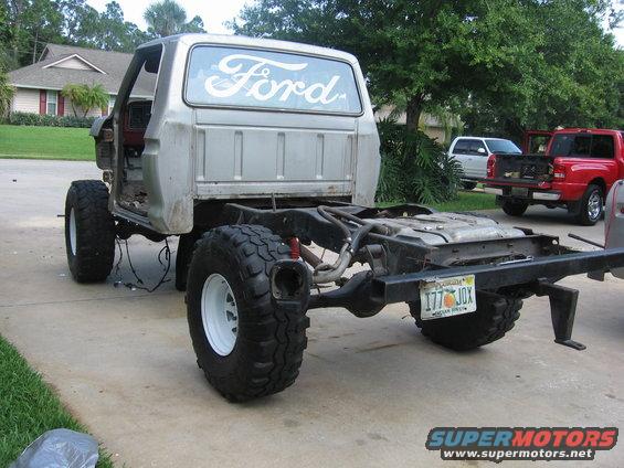 Silver Ford truck cab and chassis with large tires, missing bed and doors. White 'Ford' script on rear window