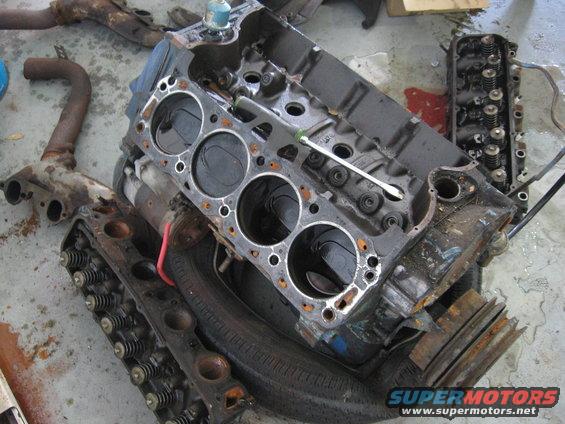 Disassembled V8 engine block and cylinder heads on a concrete floor. A screwdriver rests on the block