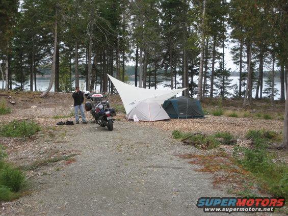 Motorcycle parked at a campsite with two tents and a tarp set up near a body of water
