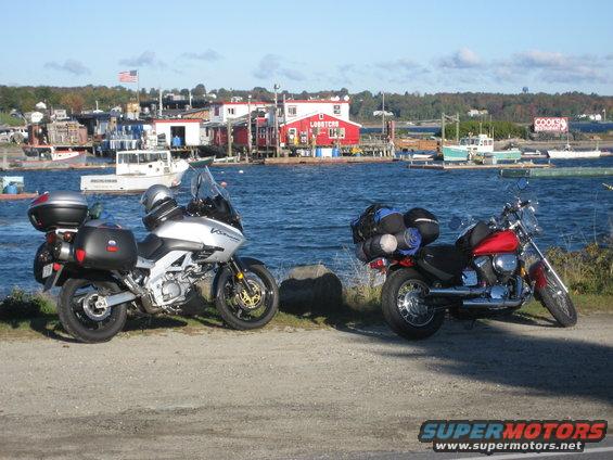 Red motorcycle parked on a gravel path with luggage on the back, facing right. A silver motorcycle is parked to its left