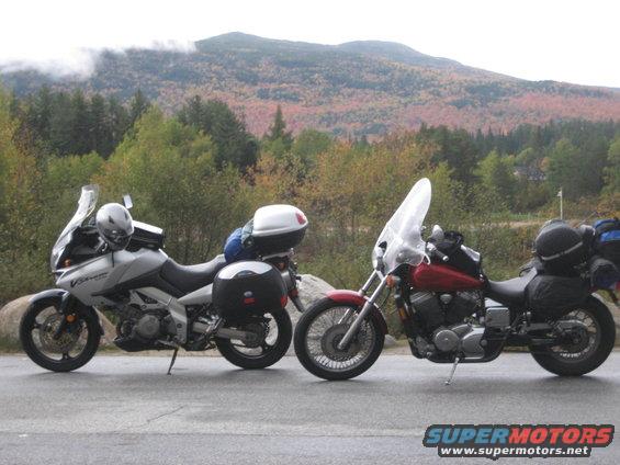 Two touring motorcycles parked on a roadside with autumn foliage and mountains in the background