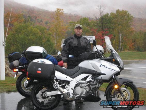 Man in riding gear stands by a silver Suzuki V-Strom motorcycle. Another motorcycle is parked behind it