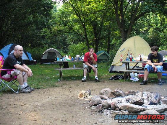 Three men sit around a campfire and picnic table at a campsite with tents in the background