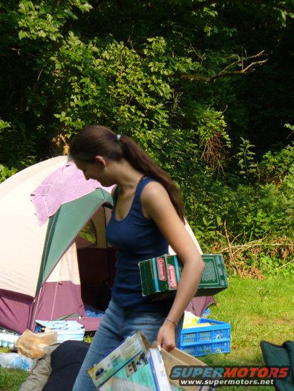 Woman in blue tank top and jeans carries boxes in front of a tent