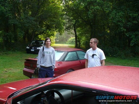 Two people stand near a red coupe, with a black sedan behind them. The red car has a spoiler