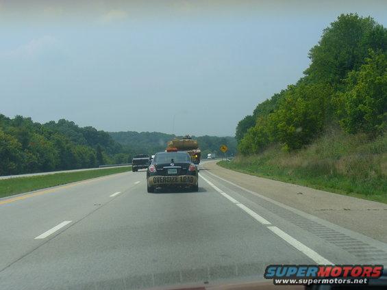 Black sedan with "OVERSIZE LOAD" tape on its rear bumper, driving on a highway