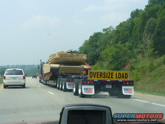 Oversize load sign on a flatbed truck carrying a tank, with a white minivan in the left lane