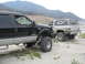 1987 Ford Bronco - Two lifted trucks, a Ford Super Duty and a Chevrolet K5 Blazer, are parked on a sandy area with mountains in the distance.