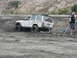 1987 Ford Bronco - A white Jeep Cherokee is stuck in thick mud, with a man observing the situation.