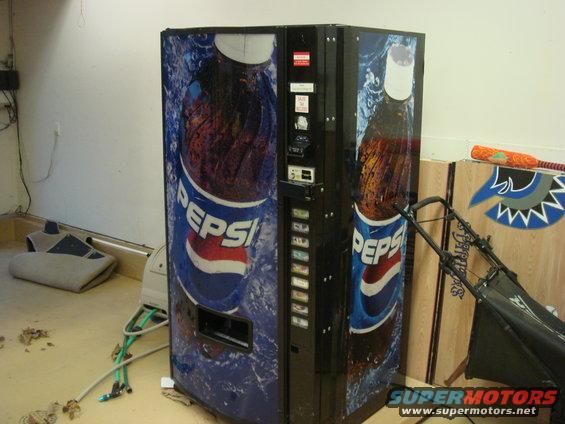 Pepsi vending machine with large Pepsi bottle graphics, next to a lawnmower and some other items