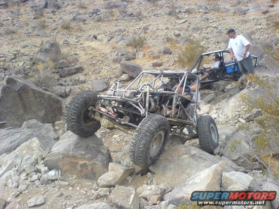 Two custom off-road buggies navigating a rocky, desert trail. One buggy is in the foreground, angled on a rock, with a person standing next to the second buggy behind it